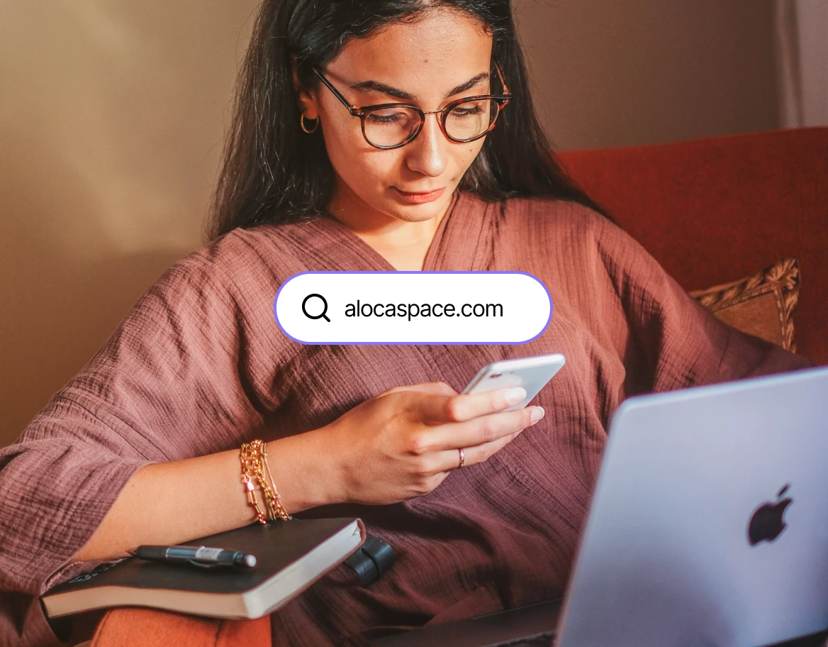 a girl looking at a phone who has laptop in hand and notebook in sofa